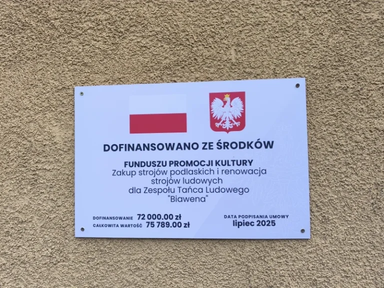 Na zdjęciu widać prostokątną tablicę informacyjną przymocowaną do chropowatej, beżowej ściany. Tablica ma białe tło, a u góry znajdują się dwa symbole: po lewej stronie flaga Polski – biało-czerwona, a po prawej godło Polski – biały orzeł w koronie na czerwonym tle. Poniżej widnieje napis: „DOFINANSOWANO ZE ŚRODKÓW FUNDUSZU PROMOCJI KULTURY” Dalszy tekst głosi: „Zakup strojów podlaskich i renowacja strojów ludowych dla Zespołu Tańca Ludowego ‘Biawena’.” Na dole tablicy umieszczono informacje finansowe: • Dofinansowanie: 72 000,00 zł • Całkowita wartość: 75 789,00 zł Po prawej stronie widnieje data: „Data podpisania umowy: lipiec 2025.” Całość utrzymana jest w przejrzystym, oficjalnym stylu, z czarnym tekstem na jasnym tle i wyraźnymi kontrastami ułatwiającymi odczyt.