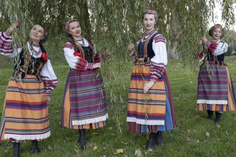 Cztery kobiety w tradycyjnych, kolorowych strojach podlaskich stoją na trawie pod drzewem, trzymając zielone witki. Uśmiechnięte, zwrócone do aparatu w ciepłym świetle popołudnia, tworzą spokojną, pogodną scenę.