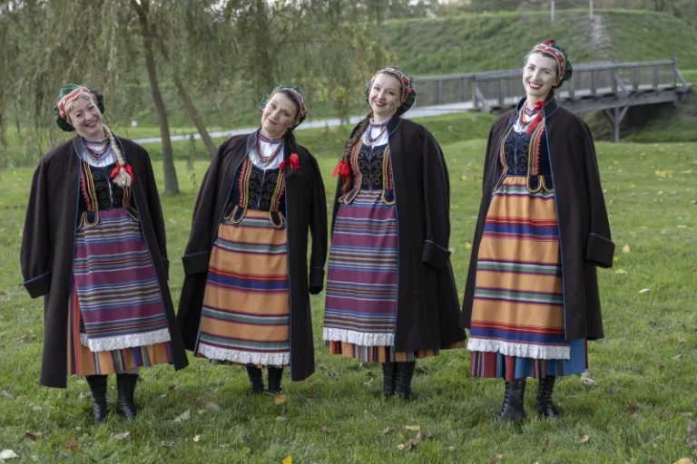 Cztery kobiety w tradycyjnych, kolorowych strojach podlaskich stoją na trawie w parku, w tle drzewa i mostek. Uśmiechnięte, lekko pochylają głowy w stronę aparatu, tworząc radosną i przyjazną scenę.