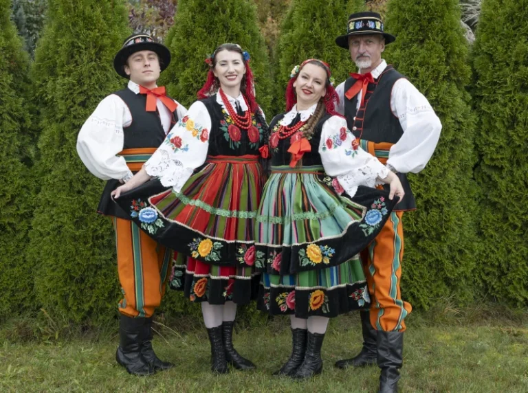 Dwie kobiety i dwóch mężczyzn w tradycyjnych, kolorowych strojach łowickich stoją parami na tle zielonych tui. Kobiety unoszą brzegi spódnic z haftami, wszyscy uśmiechają się do aparatu, tworząc barwną, radosną scenę.